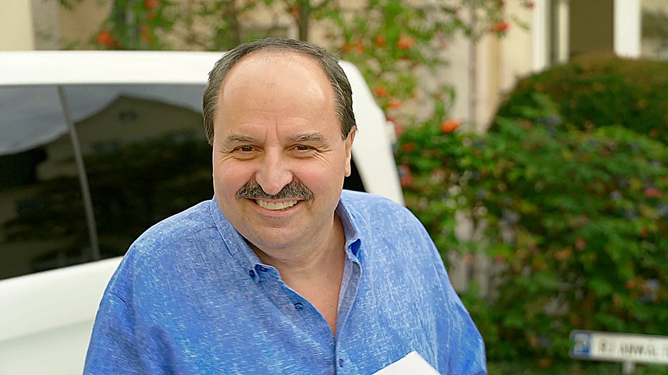 Ein Mann im blauen Hemd steht lachend vor einem weißen Fahrzeug, der Blick freundlich und offen – eine einladende Szene im Freien.