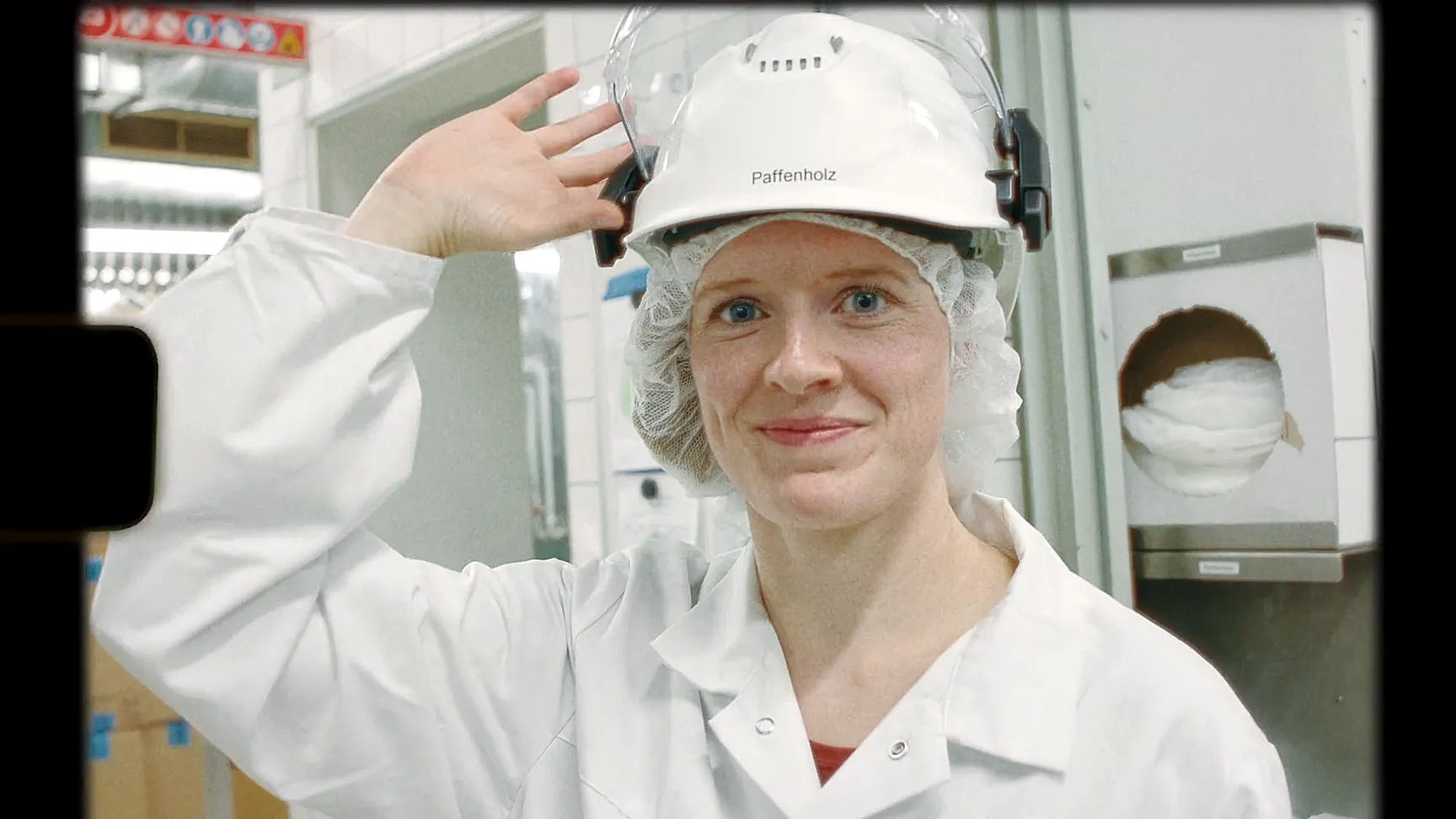 Frau in Schutzkleidung und Helm mit Gesichtsschutz, lächelnd in einer Lebensmittelproduktionsanlage.