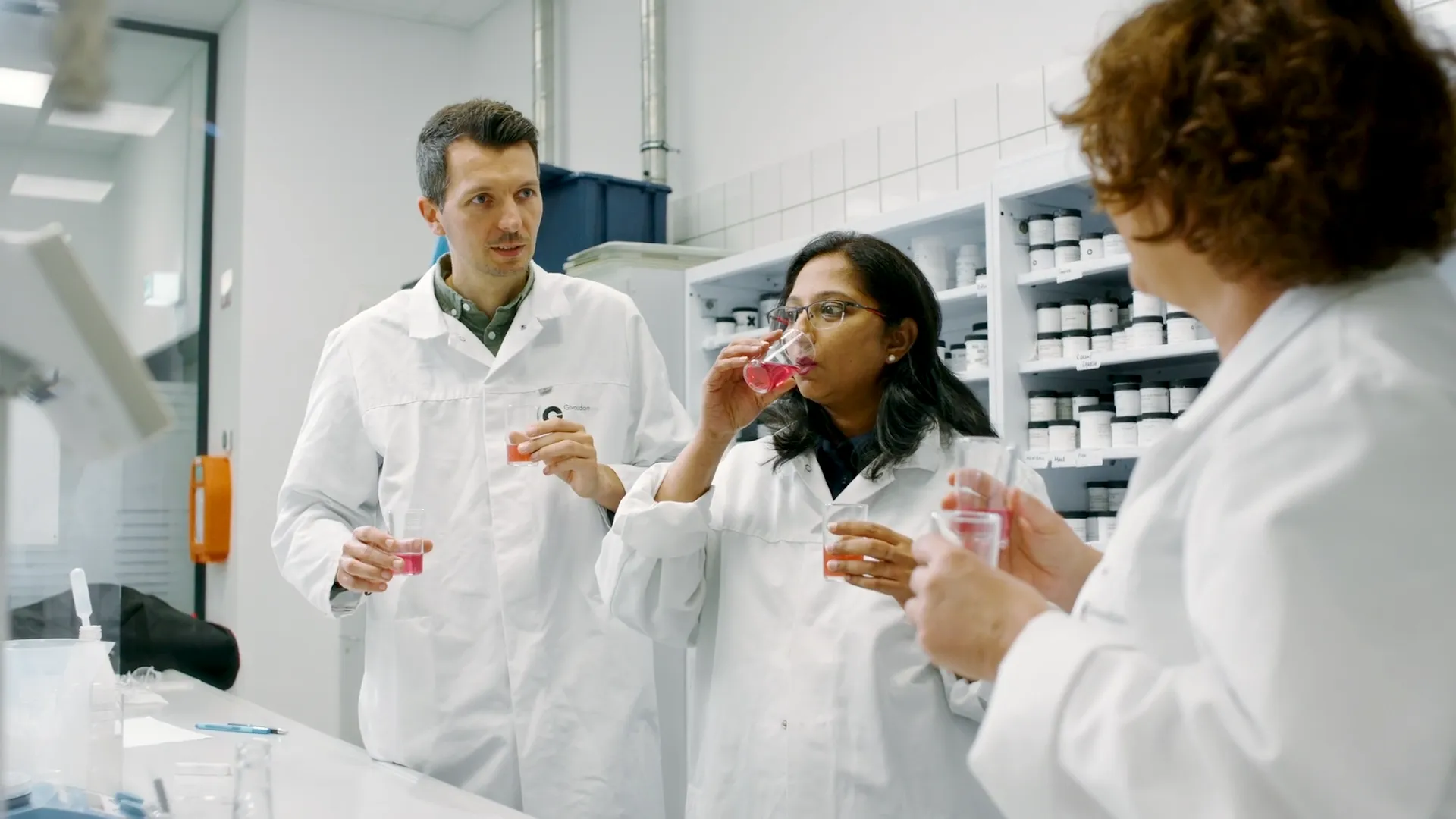 Drei Personen in weißen Laborkitteln verkosten rosa Flüssigkeit in einem Labor mit Regalen voller Proben.