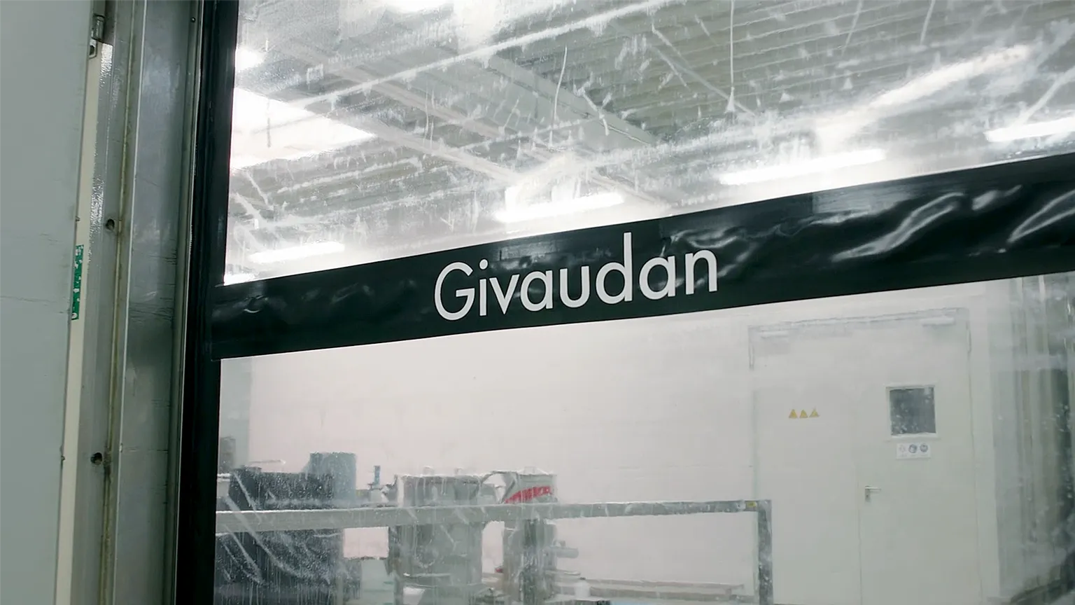 Glasfenster mit schwarzem Streifen und weißer Aufschrift ‚Givaudan‘, dahinter ein steriler Produktionsraum.