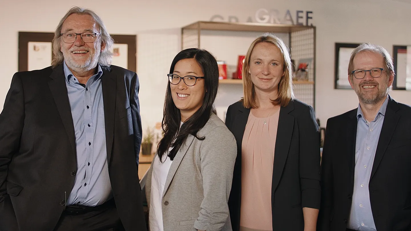 Vier Personen in Businesskleidung stehen nebeneinander, lächeln in die Kamera, im Hintergrund ist das Graef-Logo und ein Regal zu sehen.