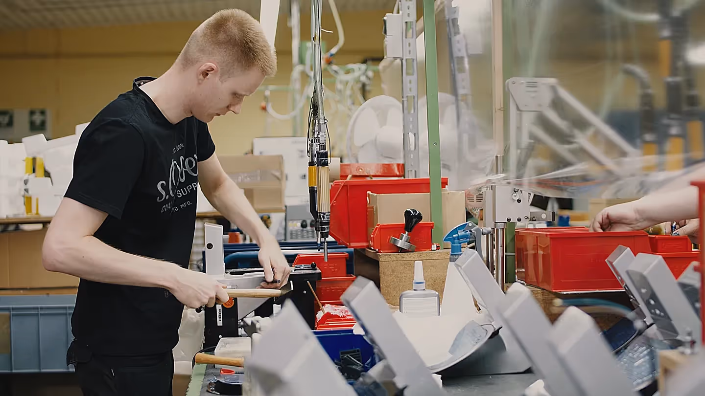 Ein Mitarbeiter in schwarzem T-Shirt arbeitet konzentriert an einer Werkbank in einer Produktionshalle, umgeben von roten Kisten, Werkzeugen und Maschinen.