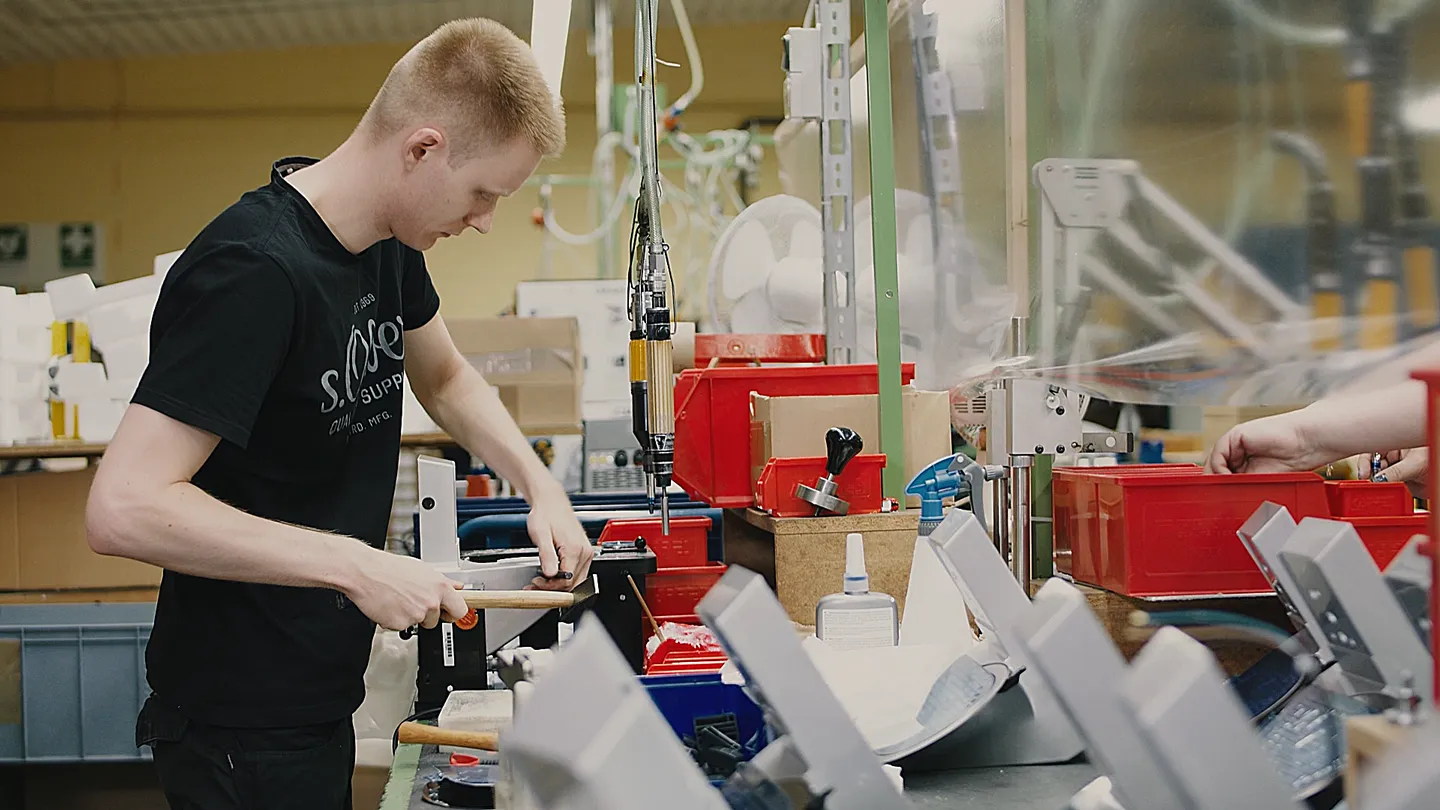Ein Mitarbeiter in schwarzem T-Shirt arbeitet konzentriert an einer Werkbank in einer Produktionshalle, umgeben von roten Kisten, Werkzeugen und Maschinen.