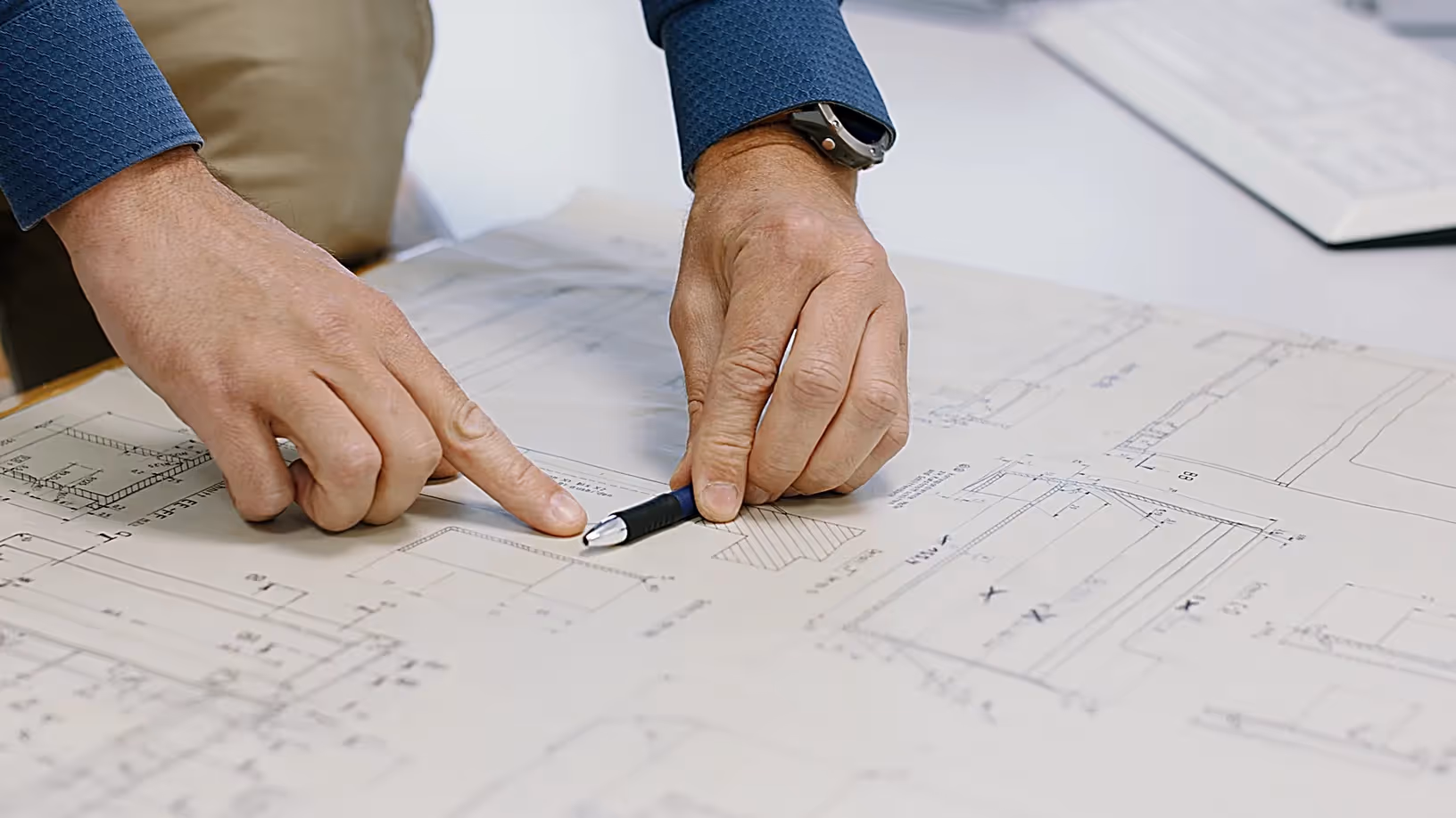 Nahaufnahme von zwei Händen, die mit einem Stift auf technische Zeichnungen und Baupläne zeigen, auf einem Tisch neben einer Tastatur.