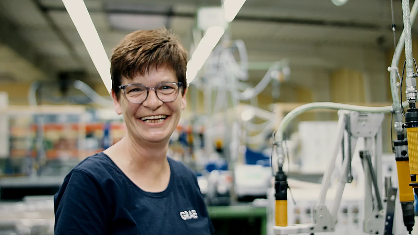 Porträt einer lachenden Mitarbeiterin in einer Produktionshalle, umgeben von Maschinen und Schläuchen, die ein freundliches und positives Arbeitsumfeld vermitteln.