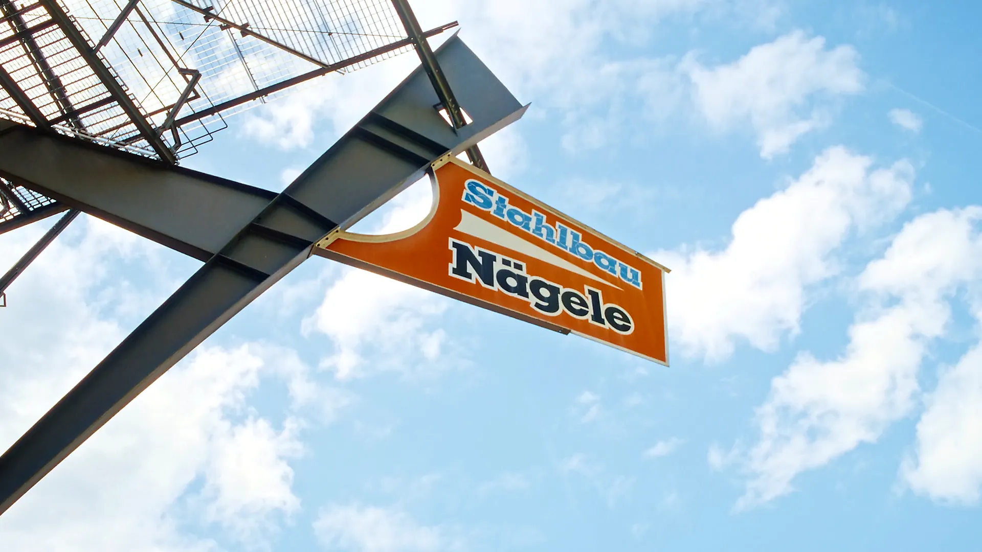 Firmenschild Stahlbau Nägele an Stahlkonstruktion vor blauem Himmel.