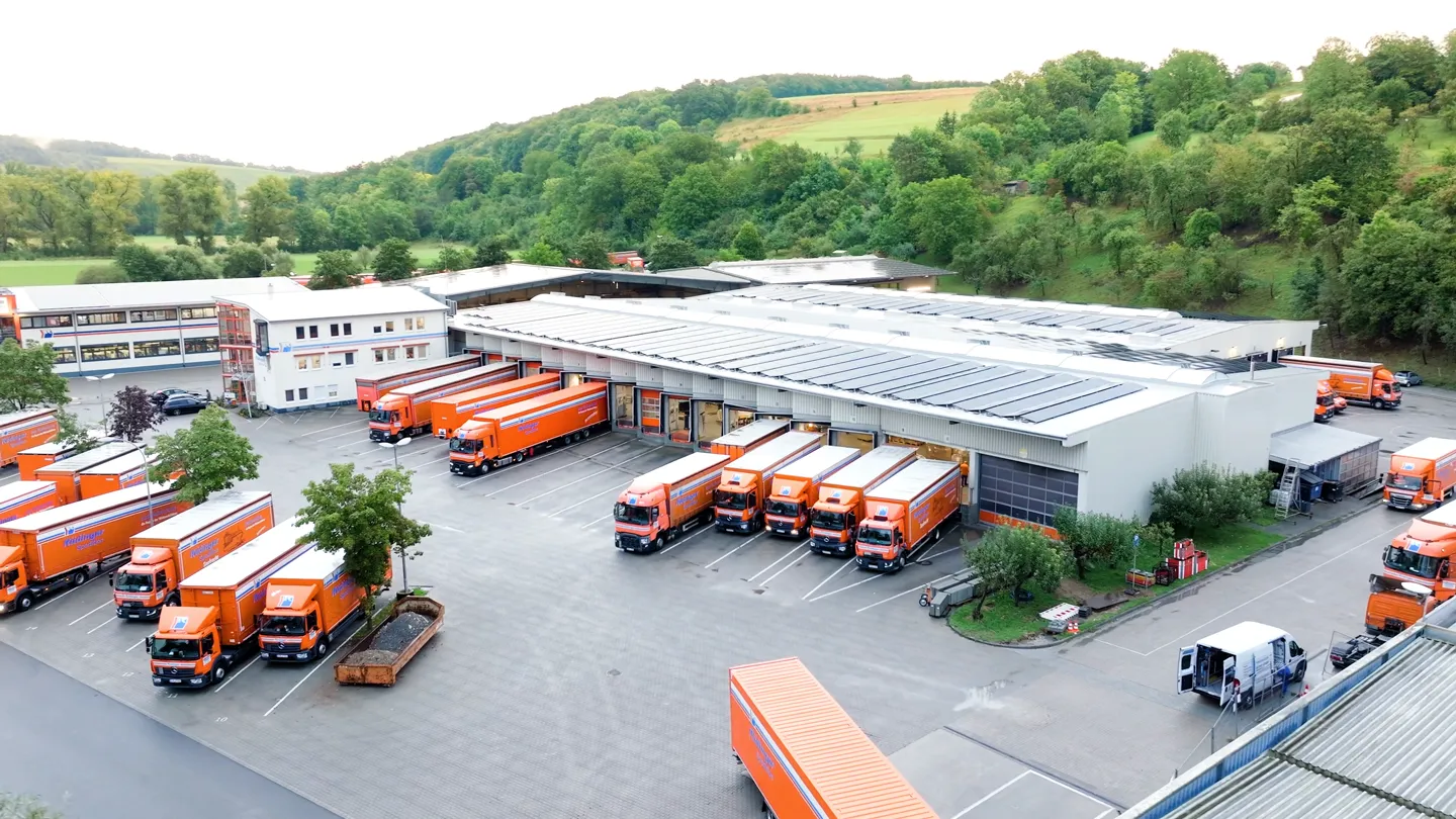 Filmproduktion Szene: Logistikzentrum mit orangefarbenen Lkw vor moderner Lagerhalle – Drohnenaufnahme zur Darstellung von Transport, Industrie und nachhaltiger Logistik in professioneller Filmproduktion.