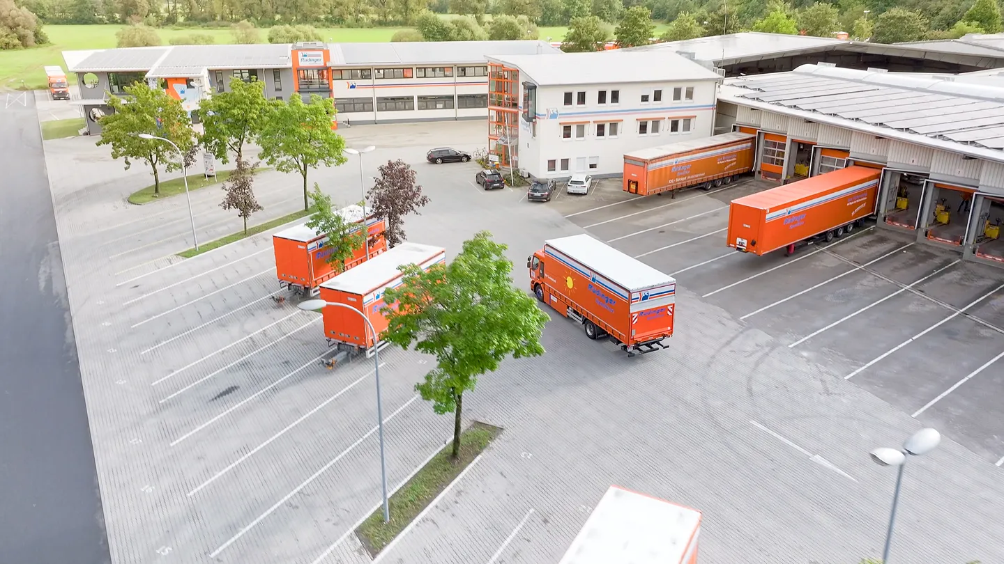 Logistikzentrum der Rüdinger Spedition mit orangefarbenen Lkw und Anhängern auf dem Hof – Drohnenaufnahme für Unternehmensfilm, Transportlogistik und moderne Industriekommunikation in professioneller Filmproduktion.