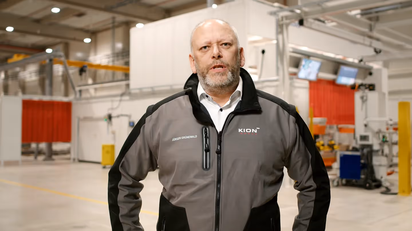 KION Produktionsleiter in moderner Fertigungshalle, Mitarbeiter trägt KION Jacke mit Logo und spricht in die Kamera.