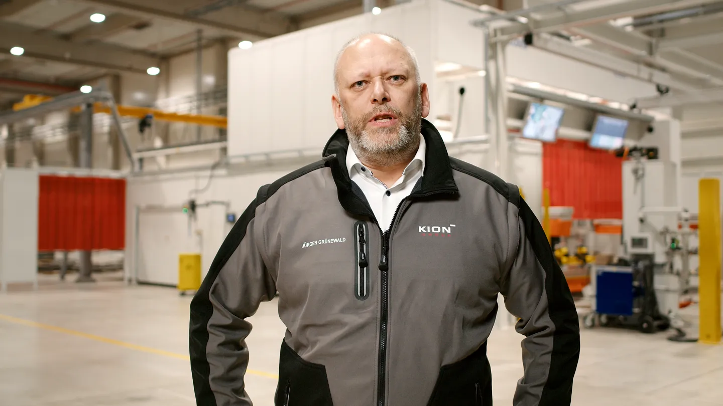 KION Produktionsleiter in moderner Fertigungshalle, Mitarbeiter trägt KION Jacke mit Logo und spricht in die Kamera.