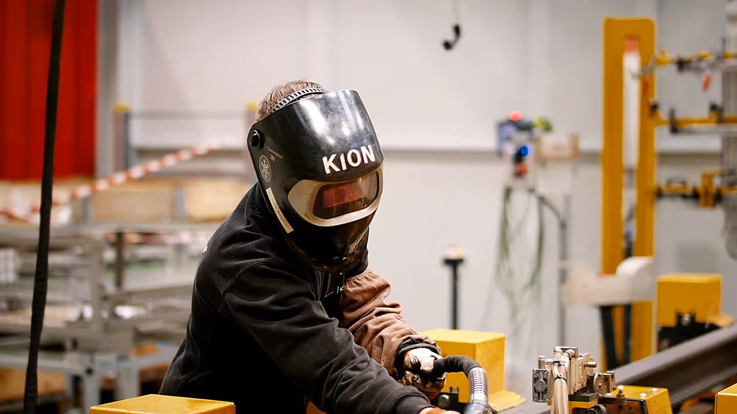 KION Mitarbeiter mit Schweißhelm arbeitet in moderner Produktionshalle an einer Metallkonstruktion.