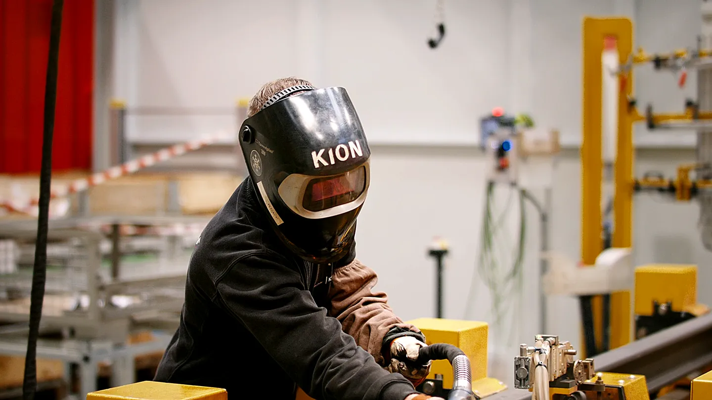 KION Mitarbeiter mit Schweißhelm arbeitet in moderner Produktionshalle an einer Metallkonstruktion.