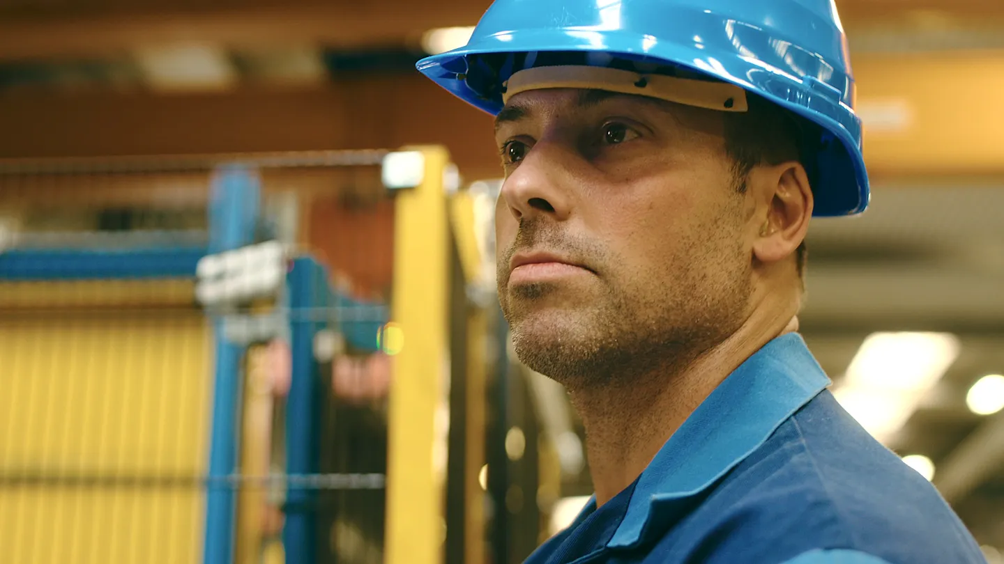Industriearbeiter mit blauem Schutzhelm und Arbeitskleidung schaut konzentriert in einer modernen Produktionshalle.