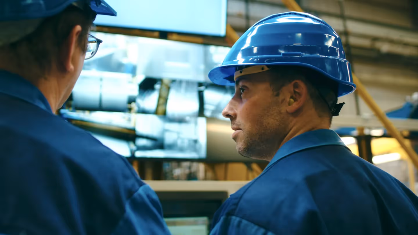 Zwei Industriearbeiter mit blauen Helmen und Schutzkleidung überwachen Produktionsprozesse an einem Monitor in einer modernen Fertigungshalle.