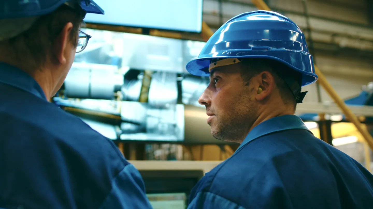 Zwei Industriearbeiter mit blauen Helmen und Schutzkleidung überwachen Produktionsprozesse an einem Monitor in einer modernen Fertigungshalle.