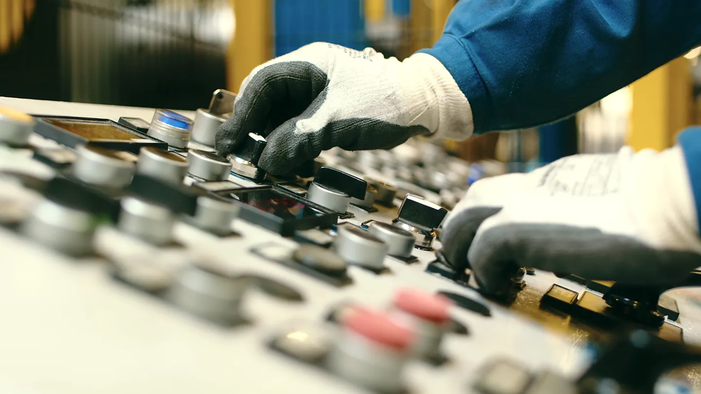 Nahaufnahme von Händen mit Arbeitshandschuhen, die eine Industriemaschine über ein Bedienfeld steuern.