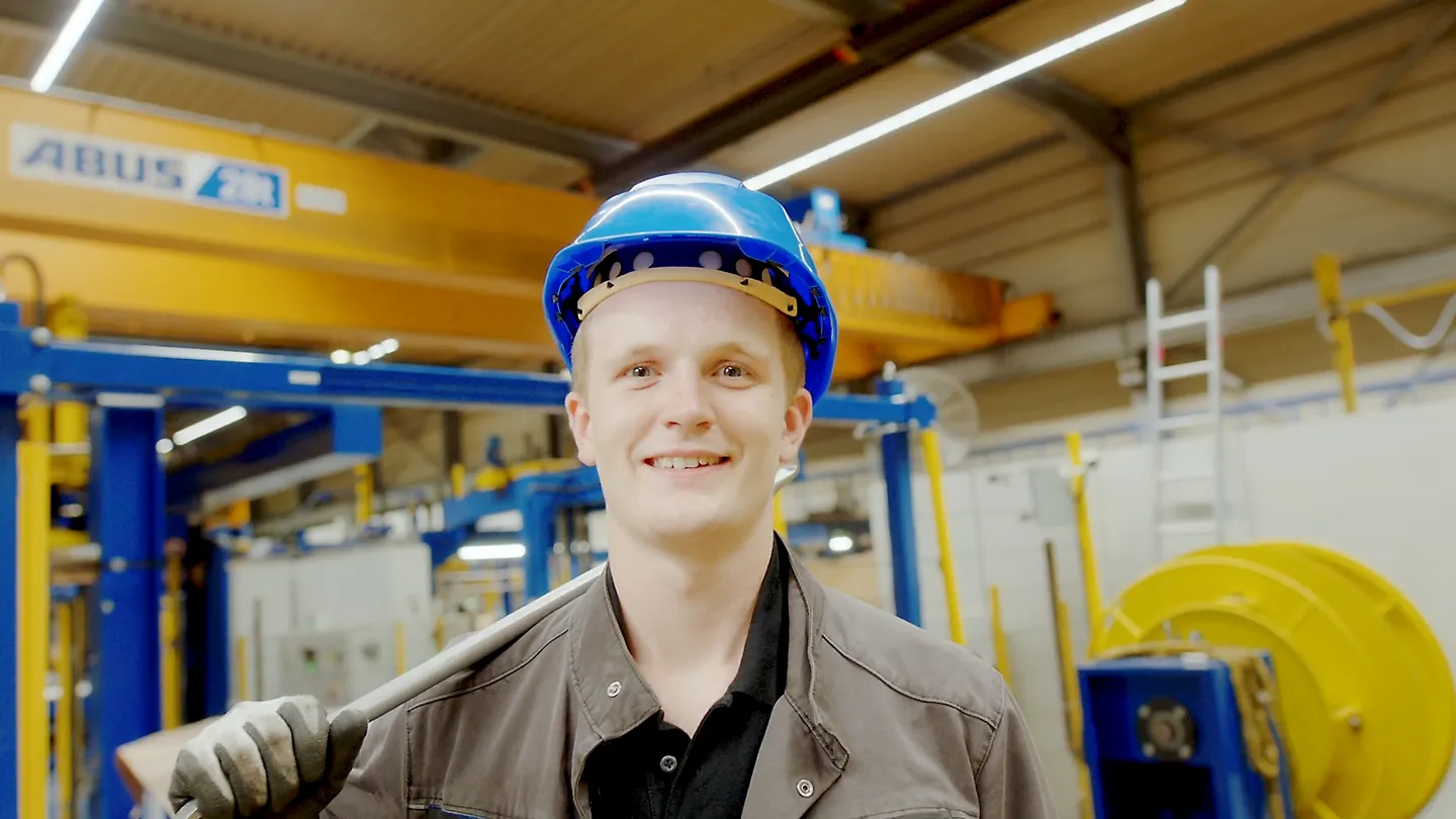 Junger Auszubildender mit blauem Helm lächelt in moderner Werkhalle, Symbol für Nachwuchs und Industrieausbildung.