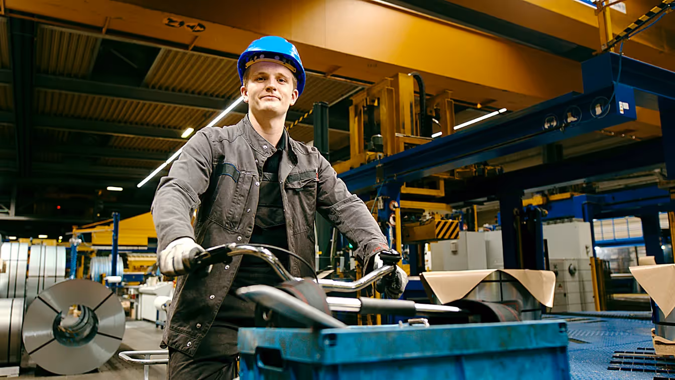 Auszubildender mit blauem Helm fährt Transportwagen durch moderne Industriehalle, Symbol für Teamarbeit und Ausbildung in der Produktion.