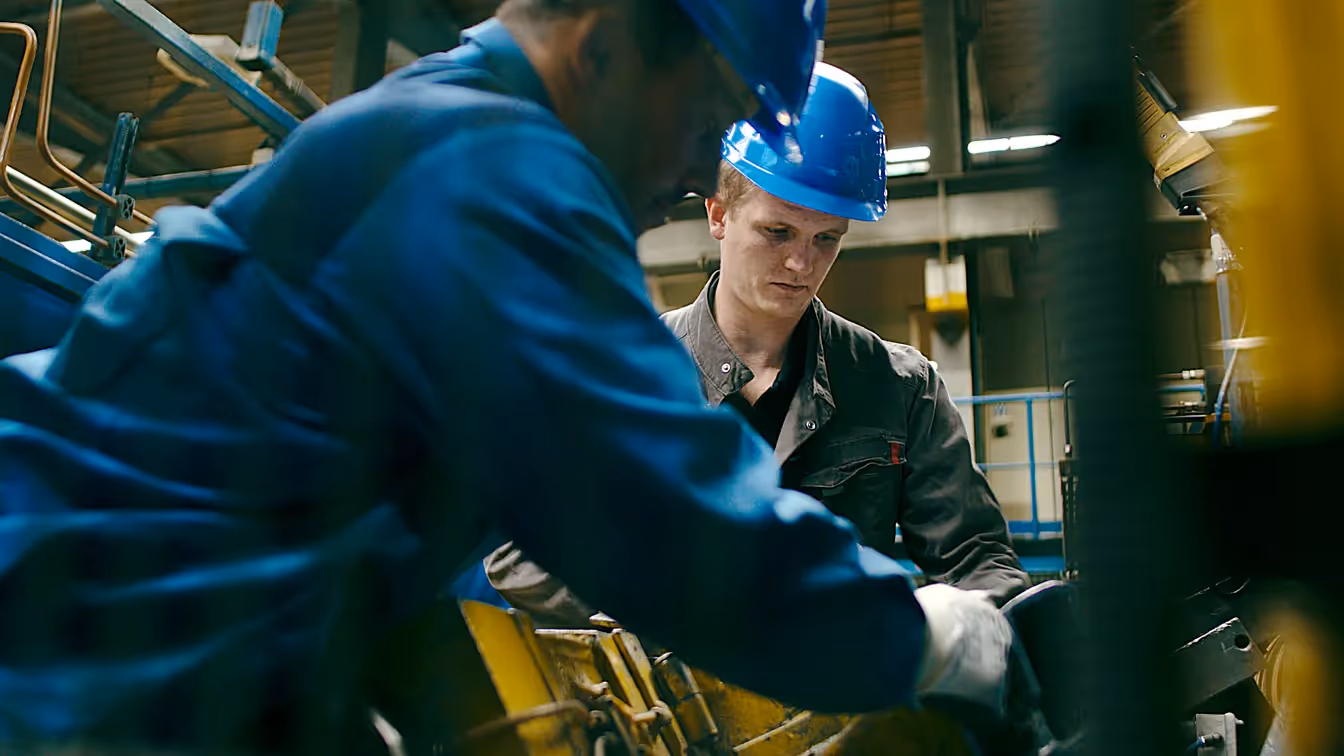 Auszubildender mit blauem Helm arbeitet konzentriert mit einem Kollegen an einer Maschine in der Produktionshalle, Sinnbild für Ausbildung, Teamarbeit und moderne Industrie.