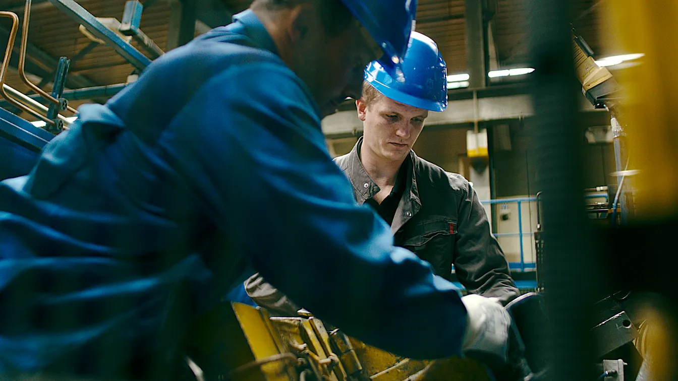 Auszubildender mit blauem Helm arbeitet konzentriert mit einem Kollegen an einer Maschine in der Produktionshalle, Sinnbild für Ausbildung, Teamarbeit und moderne Industrie.