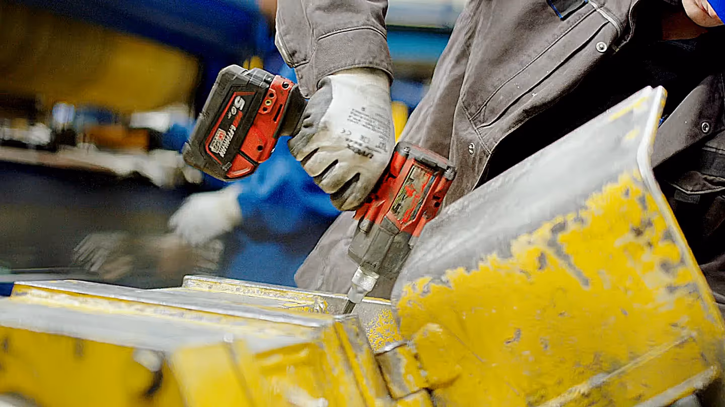 Nahaufnahme eines Auszubildenden mit blauen Helm und Arbeitshandschuhen, der mit einem roten Akkuschrauber an einer gelben Metallmaschine arbeitet – Symbol für Ausbildung, Handwerk und moderne Industrie.