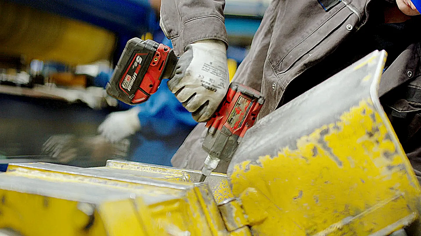 Nahaufnahme eines Auszubildenden mit blauen Helm und Arbeitshandschuhen, der mit einem roten Akkuschrauber an einer gelben Metallmaschine arbeitet – Symbol für Ausbildung, Handwerk und moderne Industrie.