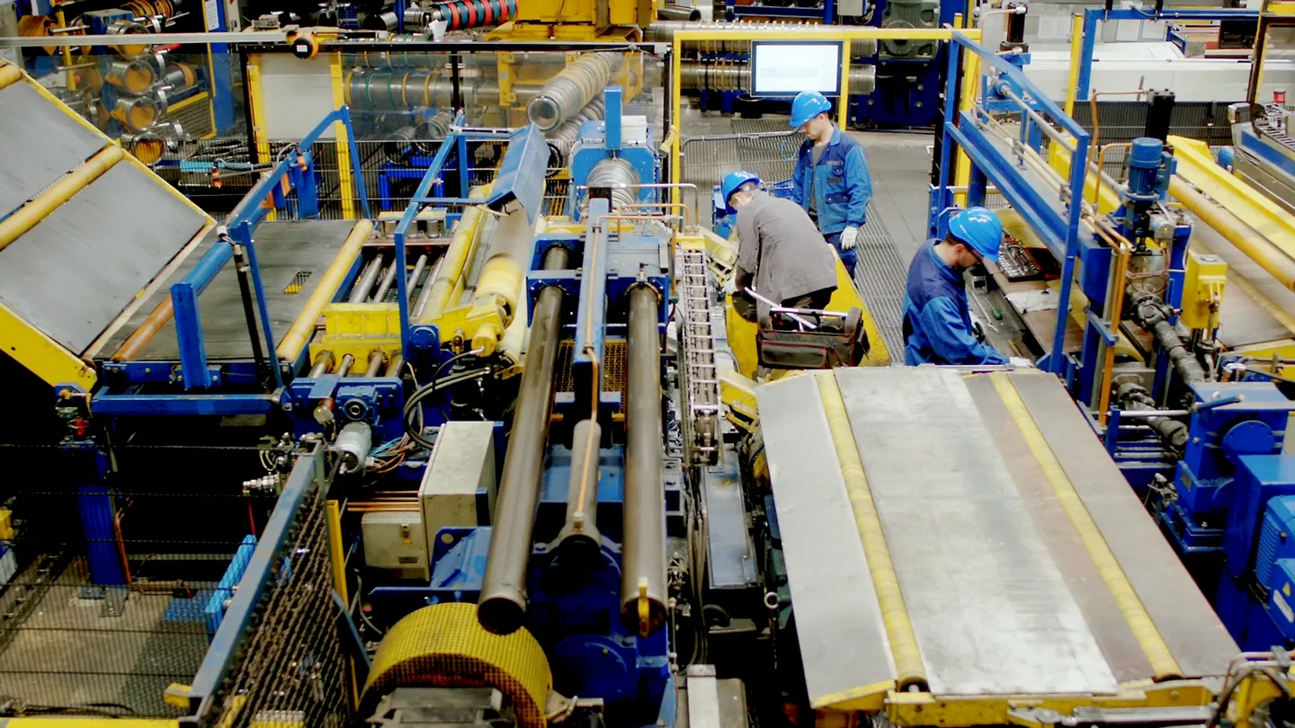 Produktionshalle mit modernen Maschinen, drei Auszubildende in blauen Helmen arbeiten an Anlagen, Symbol für Teamarbeit, Technik und Ausbildung in der Industrie.