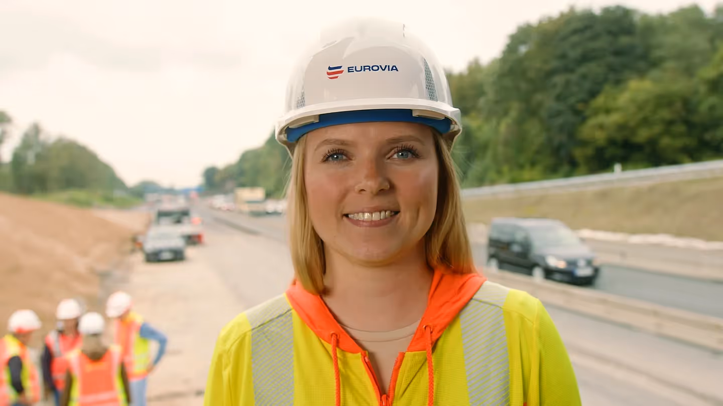 Frau mit EUROVIA-Schutzhelm und Warnjacke lächelt auf Autobahn-Baustelle, im Hintergrund Fahrzeuge und Bauarbeiter.