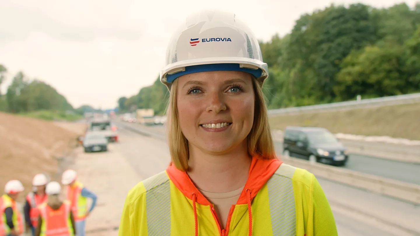 Frau mit EUROVIA-Schutzhelm und Warnjacke lächelt auf Autobahn-Baustelle, im Hintergrund Fahrzeuge und Bauarbeiter.