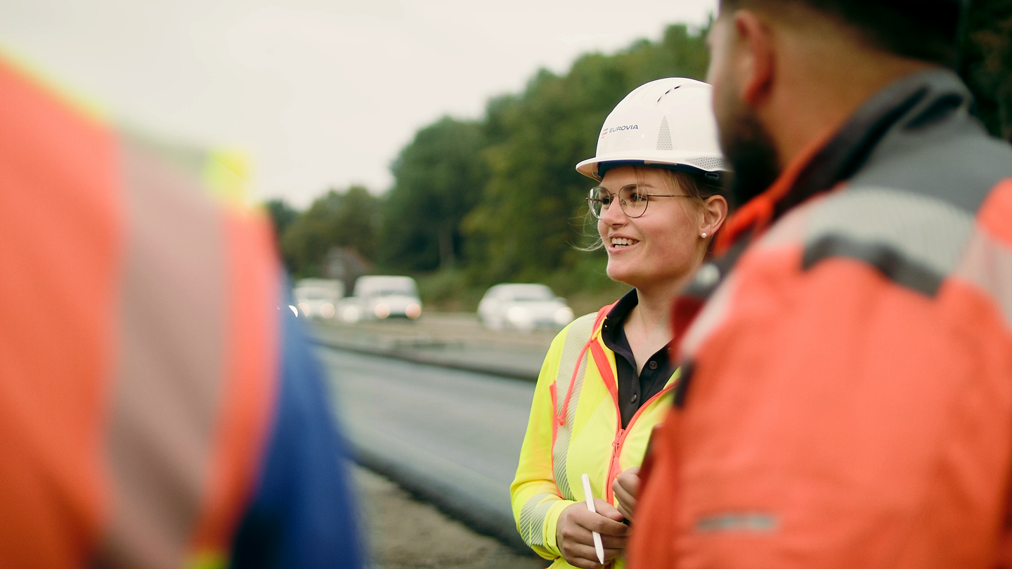 Frau mit Schutzhelm und Warnjacke lächelt im Gespräch mit zwei Kollegen auf Baustelle an einer Straße, Fahrzeuge und Bäume im Hintergrund.
