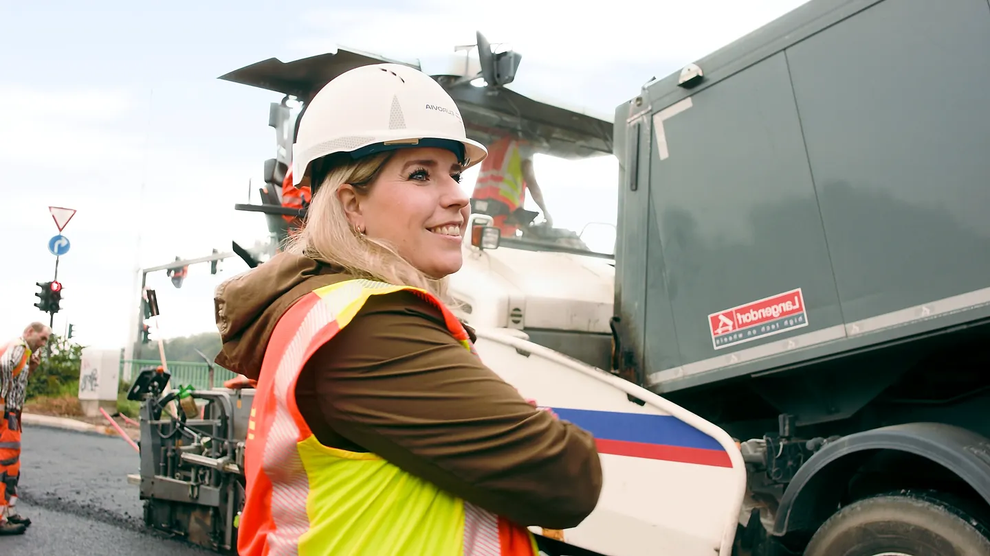 Frau mit weißem Schutzhelm und Warnweste lächelt auf Straßenbaustelle, im Hintergrund Baumaschinen und Fahrzeuge.
