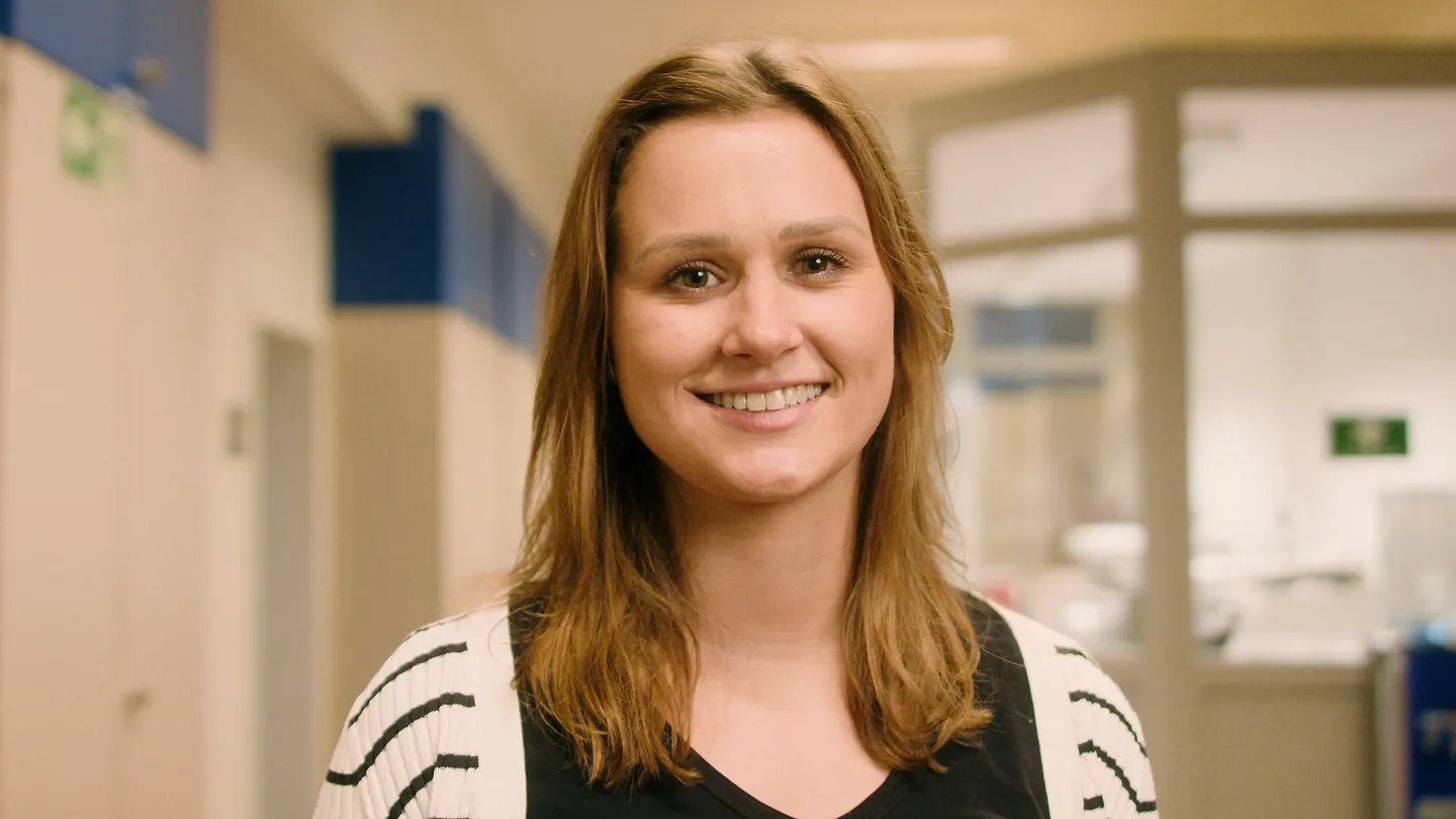 Junge Frau mit hellbraunen Haaren lächelt in hell erleuchtetem Flur, trägt schwarzes Oberteil und weiße Strickjacke mit Streifen.