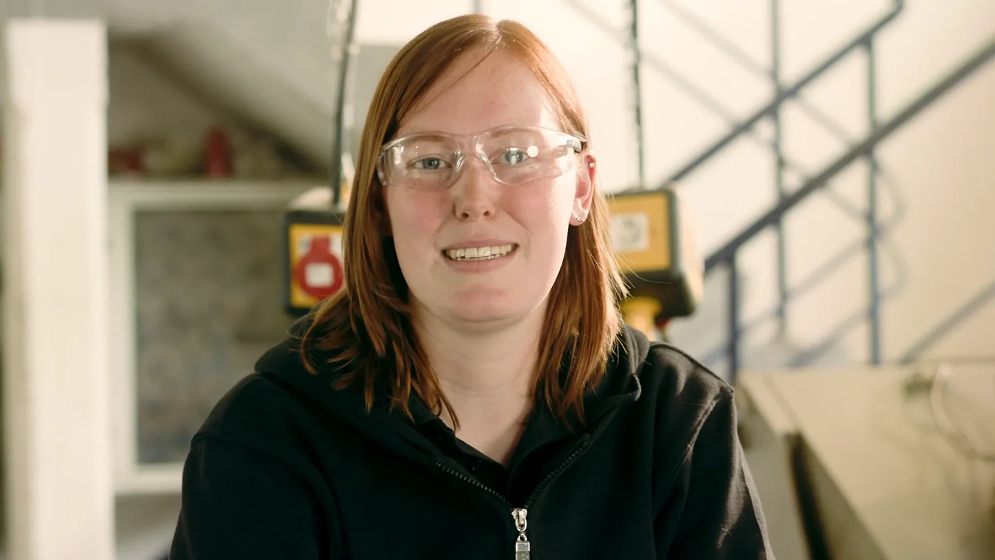 Frau mit roten Haaren, Schutzbrille und schwarzem Sweatshirt lächelt in Werkstattumgebung, unscharfe Geräte und Treppe im Hintergrund.