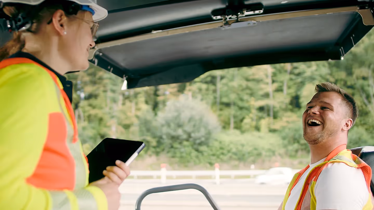 Zwei Bauarbeiter mit Schutzhelmen und Warnwesten lachen gemeinsam, einer hält ein Tablet, im Hintergrund Straße und Bäume.