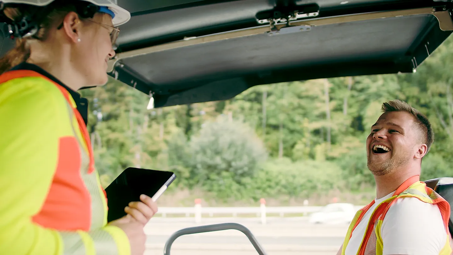 Zwei Bauarbeiter mit Schutzhelmen und Warnwesten lachen gemeinsam, einer hält ein Tablet, im Hintergrund Straße und Bäume.