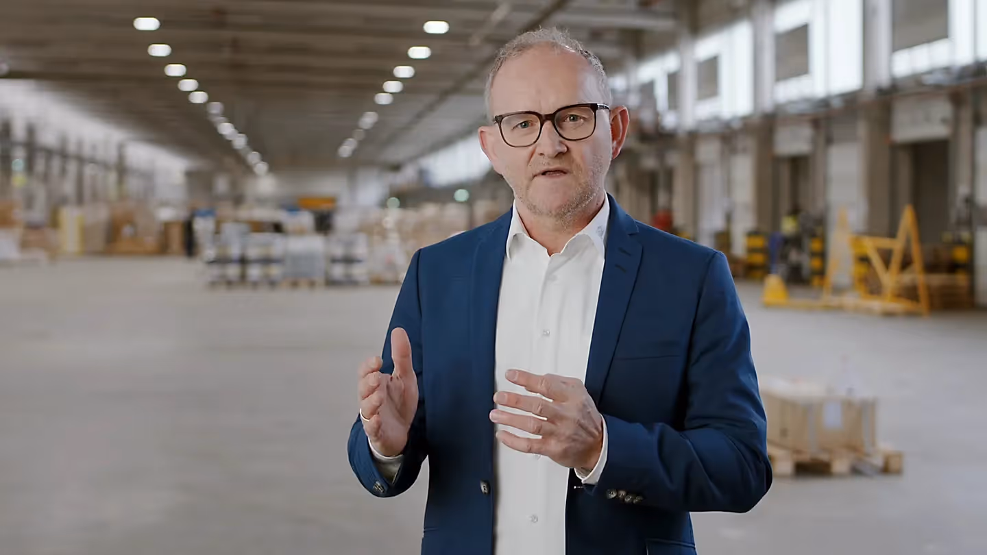 Mann im blauen Anzug spricht in einer großen Lagerhalle, Gestik mit beiden Händen.