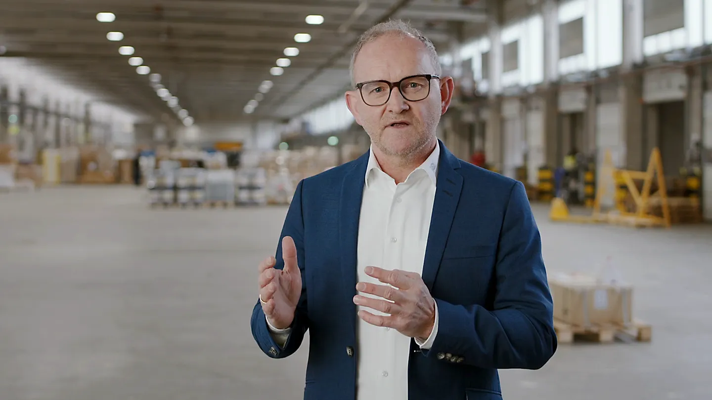 Mann im blauen Anzug spricht in einer großen Lagerhalle, Gestik mit beiden Händen.