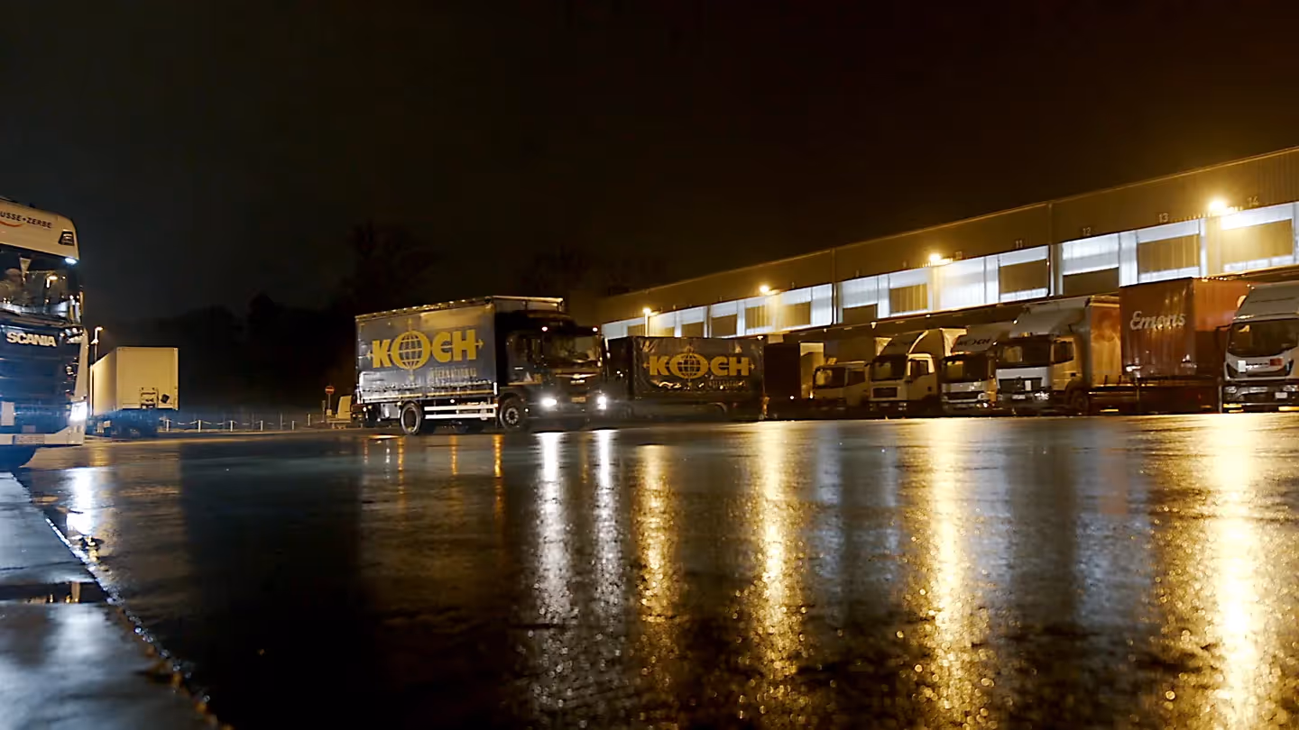 Speditionshof bei Nacht mit beleuchteten LKWs und Ladezonen, Koch Logistik LKW im Vordergrund.