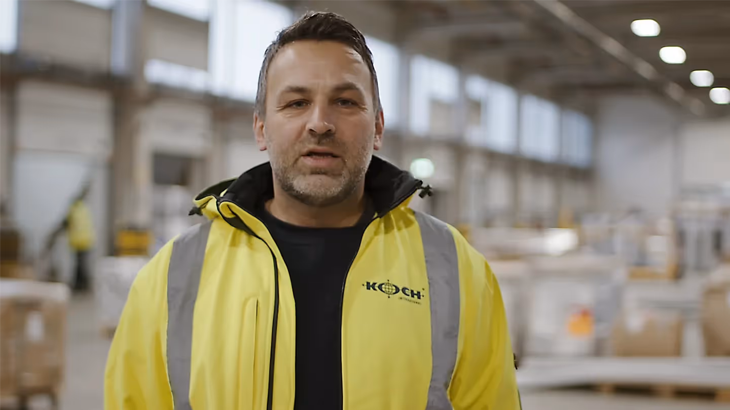 Mitarbeiter in gelber Warnjacke mit Koch International Logo spricht in Lagerhalle, Hintergrund unscharf.