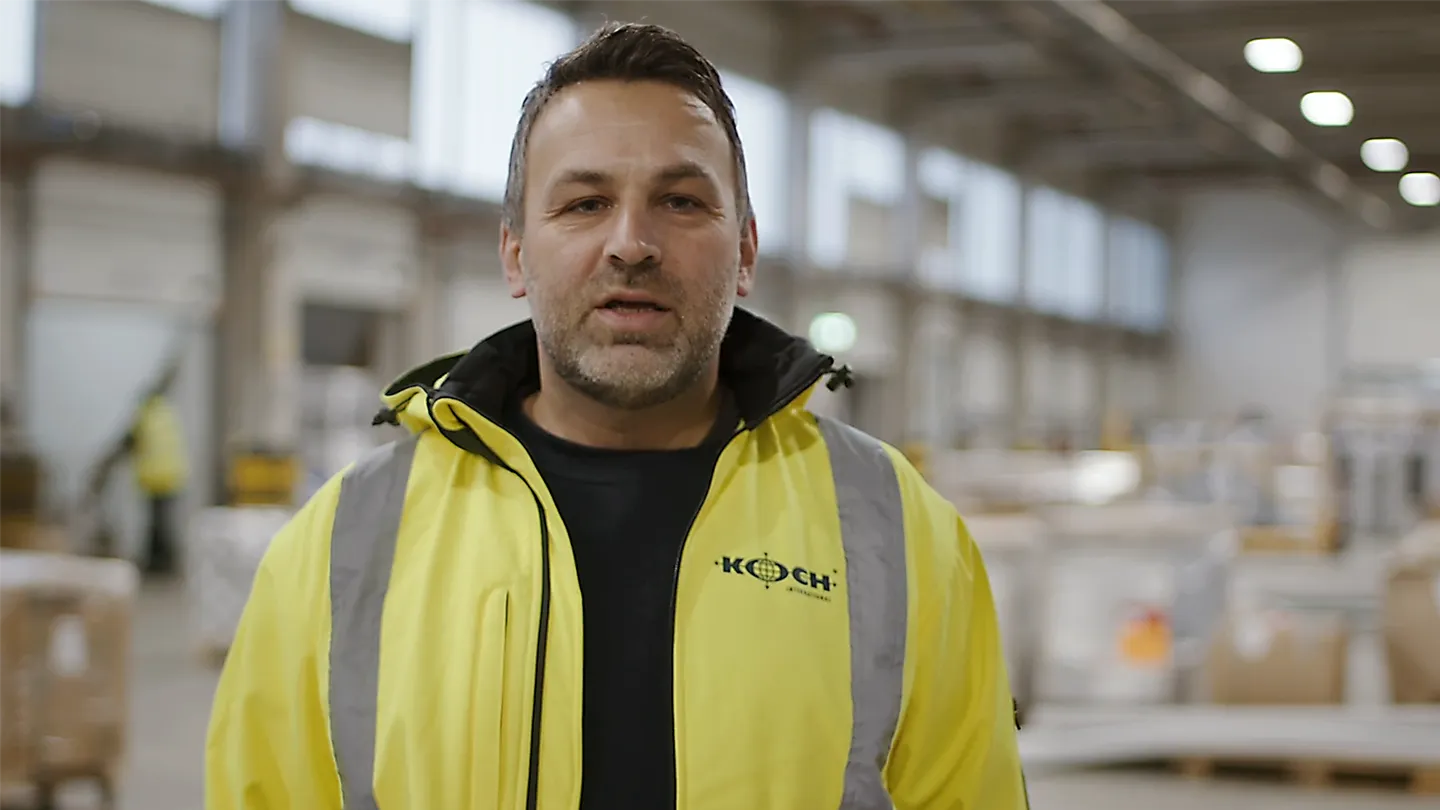 Mitarbeiter in gelber Warnjacke mit Koch International Logo spricht in Lagerhalle, Hintergrund unscharf.