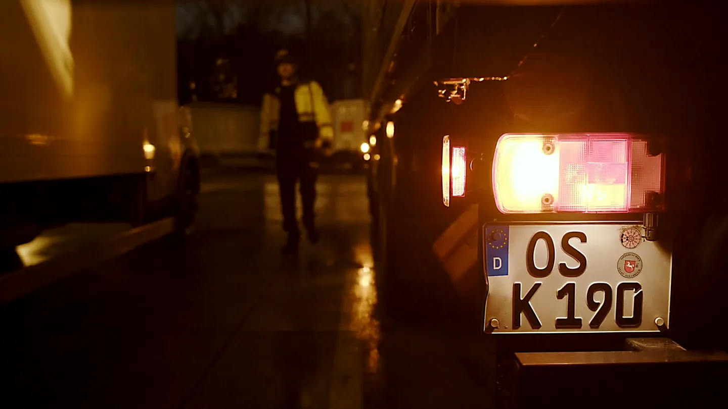 LKW Rücklicht mit leuchtendem Kennzeichen bei Nacht, Arbeiter in Warnjacke auf Speditionshof im Hintergrund.