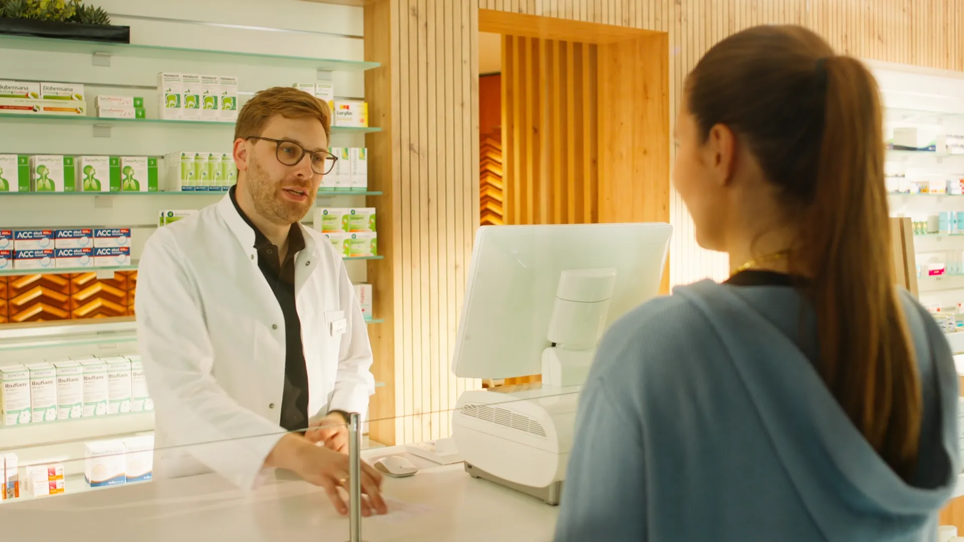 Beratungsszene in einer modernen Apotheke: Ein Apotheker im weißen Kittel spricht mit einer Kundin am Tresen. Authentisches Motiv aus einem medizinischen 2D-Erklärvideo oder Werbefilm über Gesundheit, Service und pharmazeutische Kompetenz.