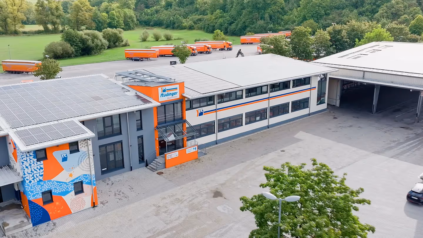 Modernes Logistik- und Industriegebäude der Rüdinger Spedition mit Solaranlage und orangefarbenen Lkw im Hintergrund – Drohnenaufnahme für Unternehmensfilm, Industrie und nachhaltige Logistik in Filmproduktion.