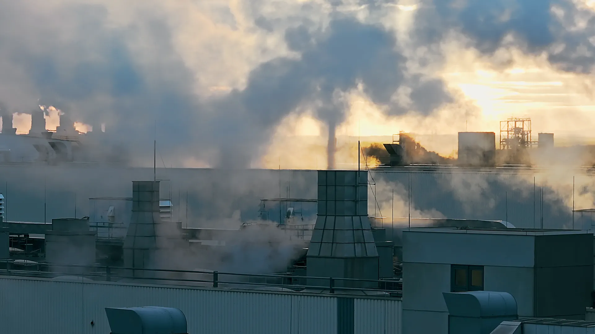 Industrieanlagen mit Rauchschwaden und Schornsteinen bei Sonnenaufgang, Symbol für Emissionen und Dekarbonisierung.