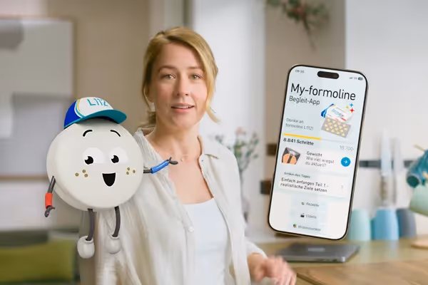 Szene aus einem Werbefilm oder Produktvideo: Frau mit Smartphone zeigt die My-formoline App, daneben das animierte Maskottchen L112 – moderne Markenkommunikation und Gesundheitsmotivation.