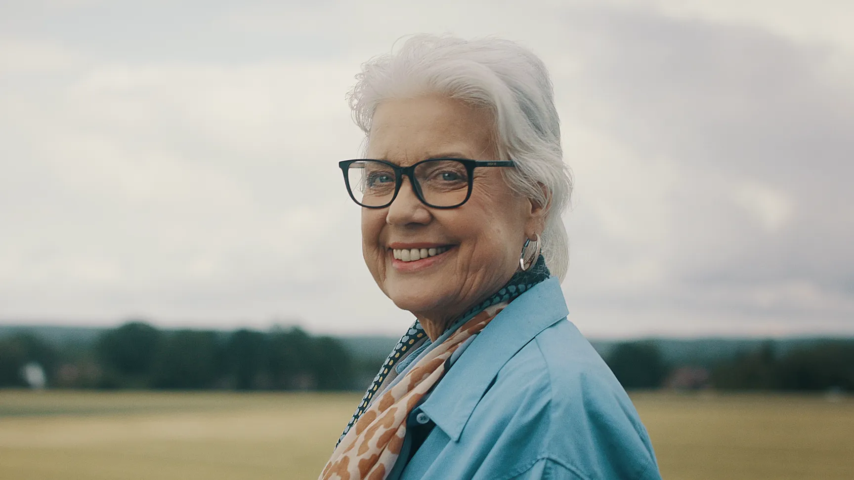 Lächelnde ältere Frau im Freien vor wolkigem Himmel. Authentische Szene für Werbefilm, Filmproduktion oder Imagevideo, die Vertrauen, Lebensfreude und menschliche Nähe vermittelt – ideal für emotionale Markenkommunikation.