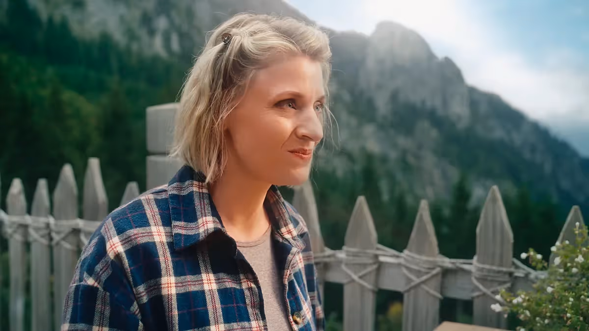 Filmproduktion mit natürlichem Licht: Frau steht vor Bergkulisse und Holzzaun, blickt nachdenklich in die Landschaft. Ruhige, authentische Szene aus einem Imagefilm mit cineastischer Bildsprache für emotionale Markenkommunikation.
