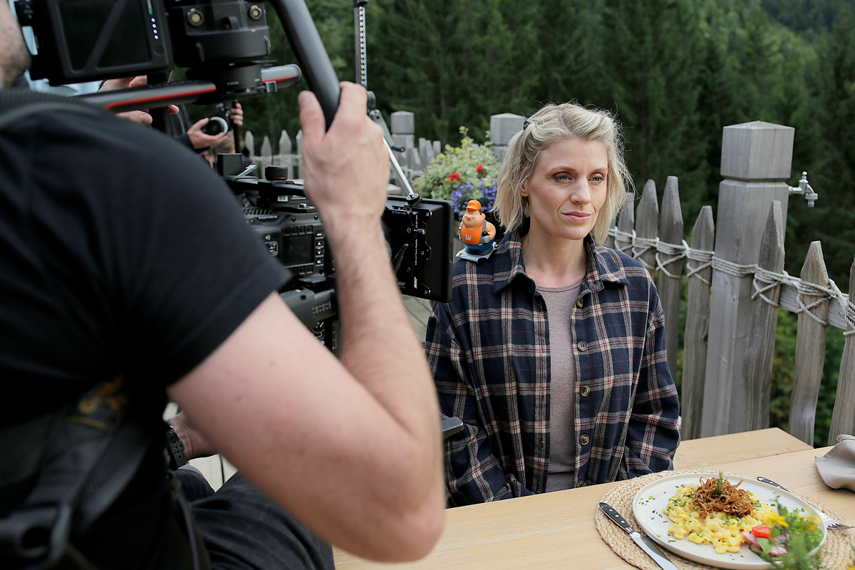 Filmszene aus einer professionellen Filmproduktion: Kameramann filmt eine Schauspielerin am gedeckten Tisch im Außenbereich. Authentische Drehsituation mit Kamera-Setup, Licht und Crew – Realfilm-Produktion für Imagefilm und Markenkommunikation.