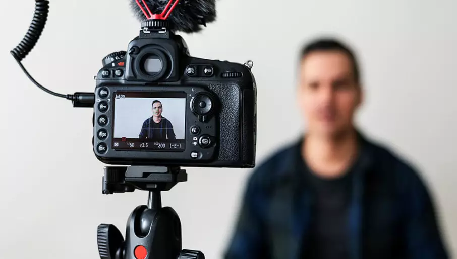 A DSLR camera is mounted on a tripod, focusing on a man whose image is displayed in sharp focus on the camera's screen. The man himself is out of focus in the background, indicating the camera's perspective is the main subject. There's a microphone attached to the camera, suggesting this setup is for video recording, possibly for a vlog or interview.