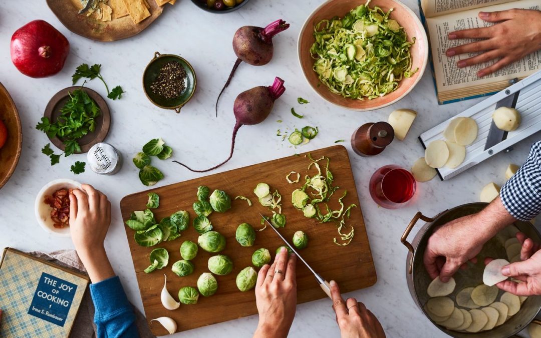 Two people are preparing vegetables; one slices potatoes with a mandoline, the other chops Brussels sprouts. A cookbook, fresh herbs, and various vegetables are visible, indicating a meal in progress.